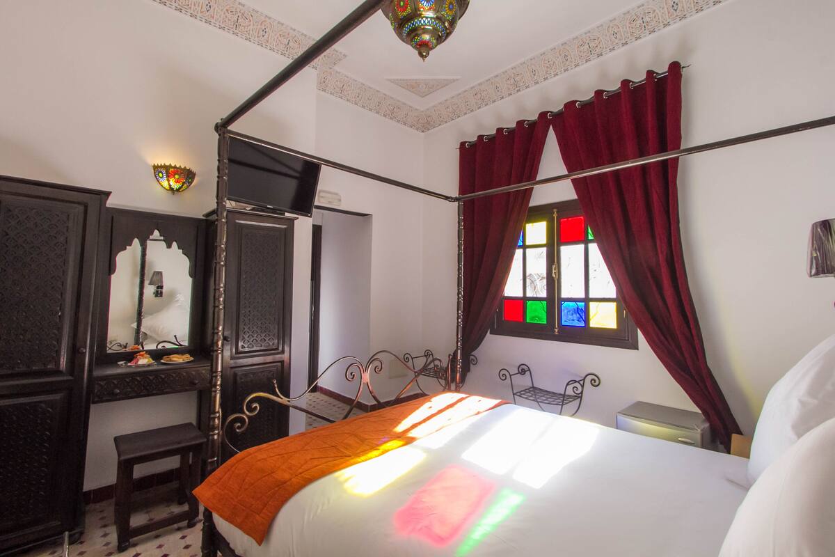 Uma cama com dossel e cortinas, mesa de cabeceira, espelho e janela.