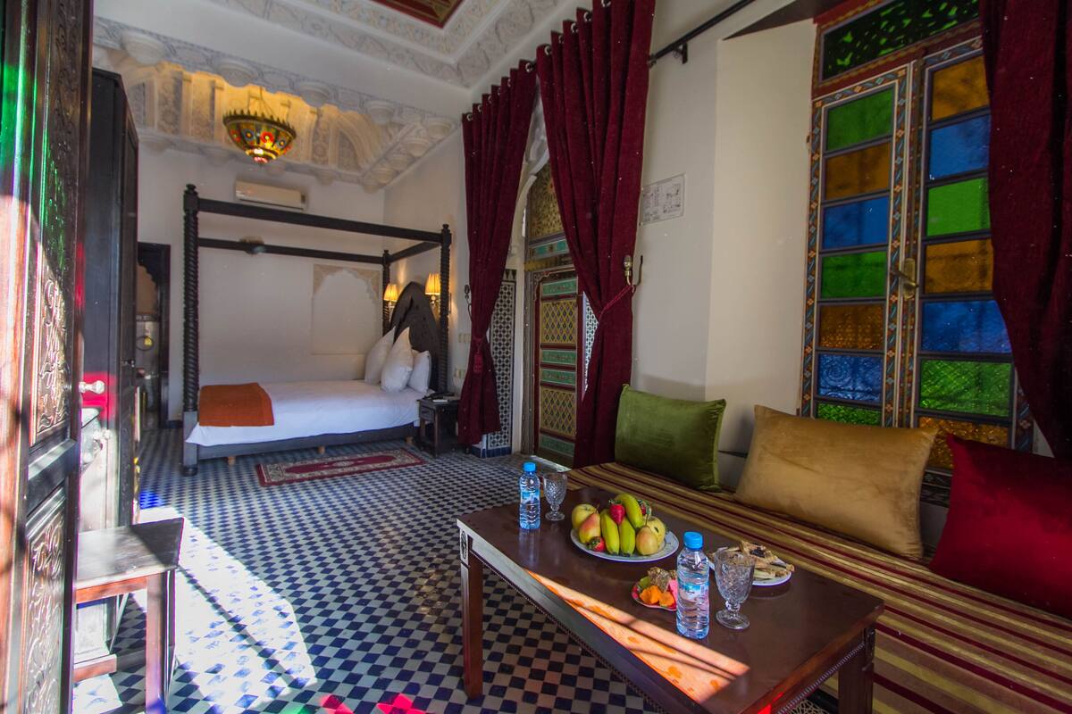 Um quarto com piso xadrez, uma cama, um sofá, uma mesa com frutas e garrafas, e uma janela com vidros coloridos.