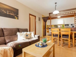 A cozy living room with a brown sofa, a coffee table with decorative items, a dining area with wooden chairs and a table, and a kitchen in the background.