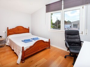 Een slaapkamer met een houten bed, een zwarte bureaustoel en een raam met uitzicht op gebouwen en groen.