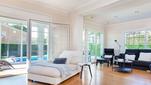 Un salon spacieux avec un grand lit, un canapé sombre et une petite table. De grandes fenêtres offrent une vue sur une piscine et sur la verdure environnante.