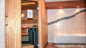 A sauna room with a wooden bench, a bench with a towel, a sauna heater, and a tiled wall with a mosaic border.