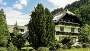 A two-story house with a sloped roof, surrounded by lush greenery and a forested mountain backdrop.
