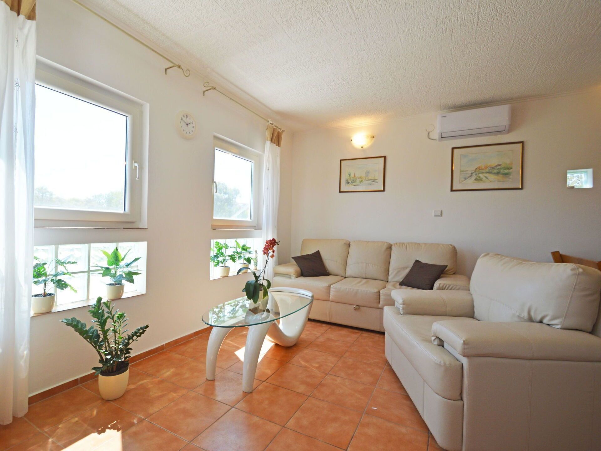 A living room with a beige sofa, a glass coffee table, and potted plants. There are two windows with curtains, a wall clock, and two framed pictures.