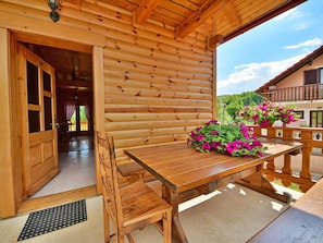 A wooden log cabin with a wooden table and chairs, a hanging flower basket, and a view of the surrounding landscape.