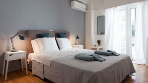 Un lit bien fait, recouvert d’une couvre-lit grise, avec deux oreillers et une lampe de chevet posée sur une table de nuit blanche.