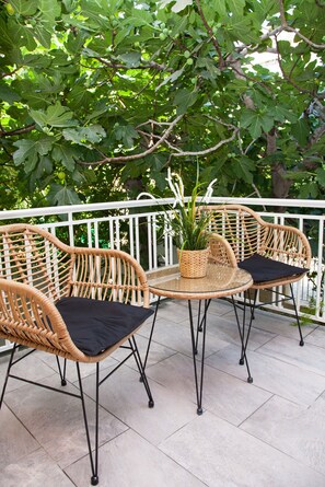 Un espace de détente extérieur aménagé avec des meubles en osier, comprenant deux chaises et une table, le tout entouré d’une rambarde blanche et d’un arbre aux feuilles vertes.