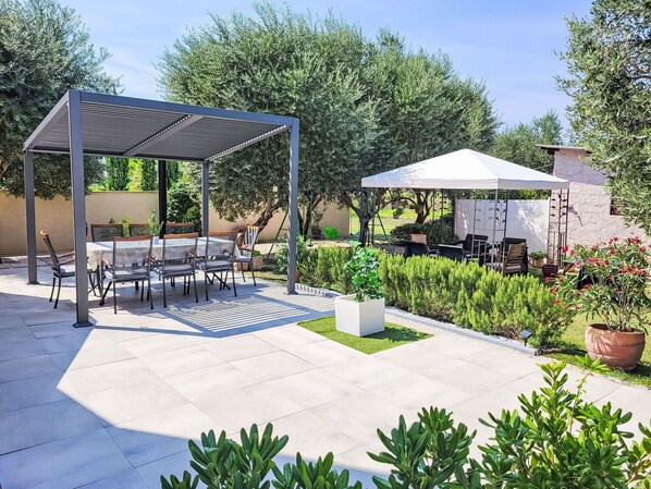 A covered patio area with a table and chairs, surrounded by greenery and a white canopy.