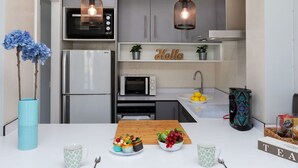 A modern kitchen with a refrigerator, microwave, and stove. There are hanging lights, a sink with lemons, and a 'Hello' sign on the shelf.