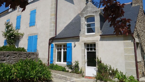 Un bâtiment à deux étages, doté de volets bleus, d’une cheminée en pierre et d’un petit porche avec une porte.