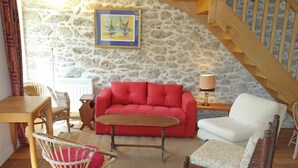 Un salon chaleureux avec un mur en pierre, un canapé rouge, un fauteuil blanc, une chaise en osier et une table en bois.