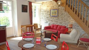 Un coin repas avec une table en bois dressée pour quatre personnes, entourée de chaises rouges. Un canapé rouge et un fauteuil blanc sont placés contre un mur en pierre. Un escalier avec des rampes en bois est visible au fond.