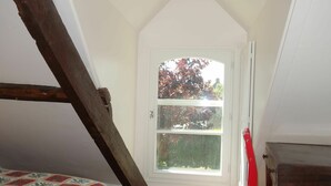 Une petite chambre mansardée avec un plafond en pente, une fenêtre donnant sur les arbres, une chaise rouge et un lit recouvert d’une courtepointe à motifs.