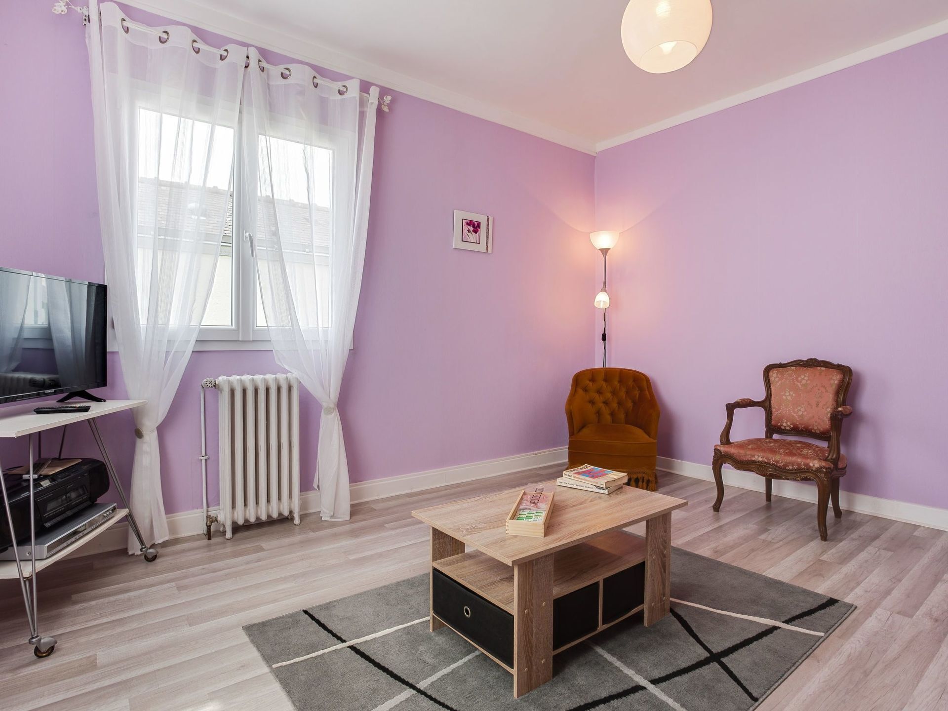 Ein Zimmer mit Fernseher auf einem Schrank, einem Heizkörper, einem Tisch mit Büchern, zwei Stühlen und einer Lampe.