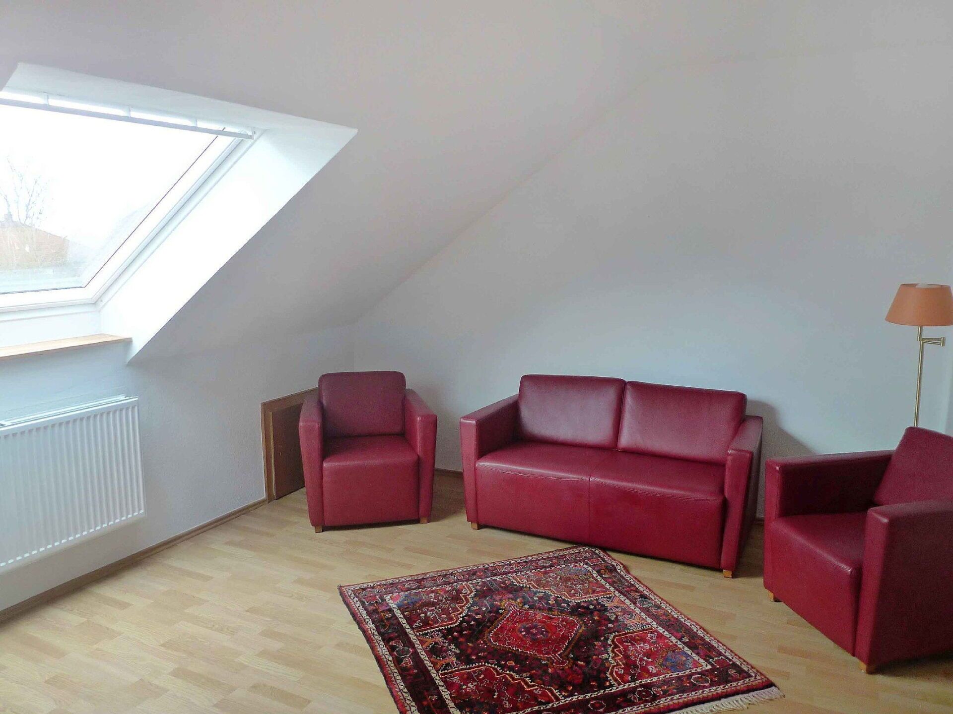 A room with a sloped ceiling, a red sofa, a smaller red armchair, a patterned rug, and a floor lamp.