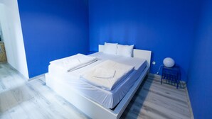 Un dormitorio con una cama, ropa de cama blanca, una pared azul y una mesita con un objeto blanco.