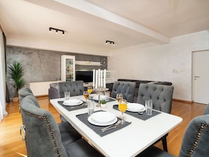 A modern dining area with a white table set for four, grey upholstered chairs, a buffet cabinet, and a television in the background.