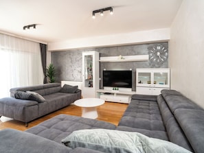 A modern living room with grey sofas, a flat-screen TV, a glass cabinet, and a clock on the wall.