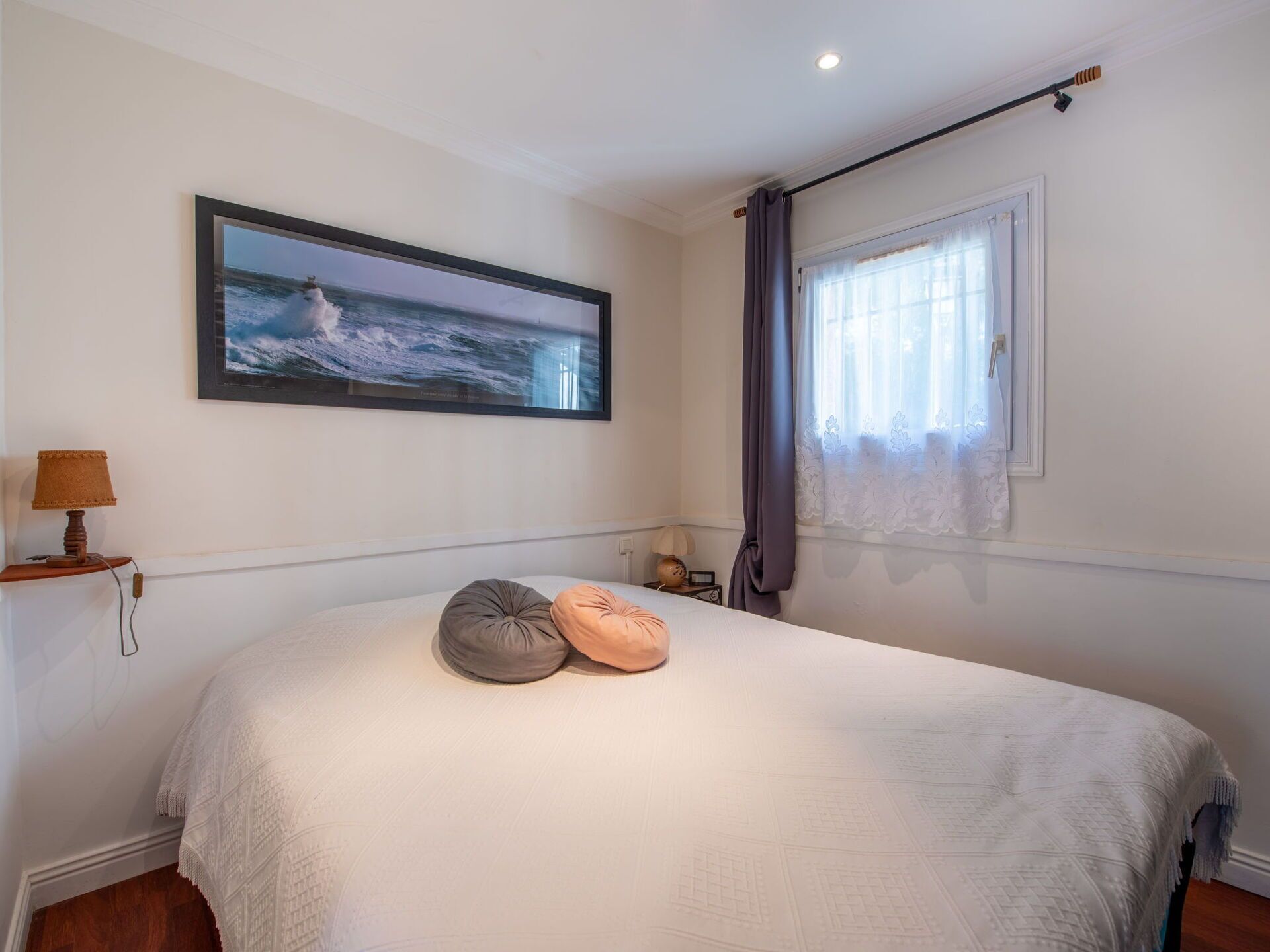 A bedroom with a large bed, a framed picture of the ocean, a lamp, and a window with curtains.