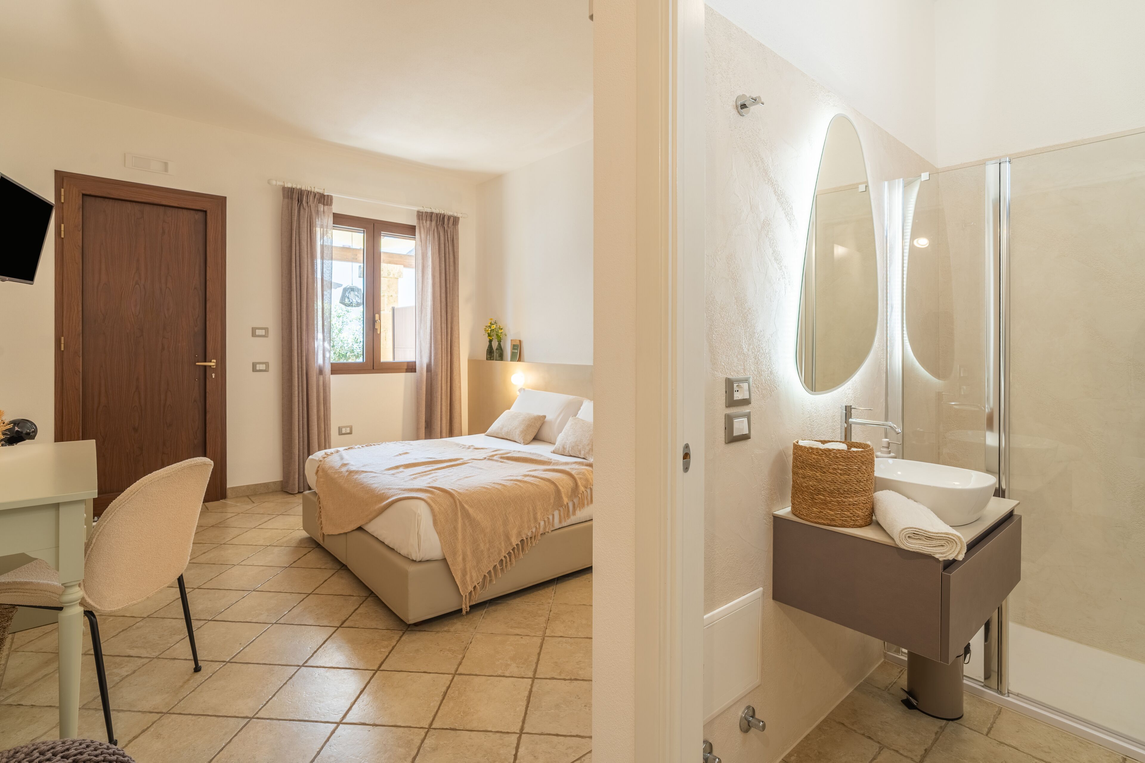 Un dormitorio con cama, escritorio, silla, ventana con cortinas y baño con lavamanos y espejo.