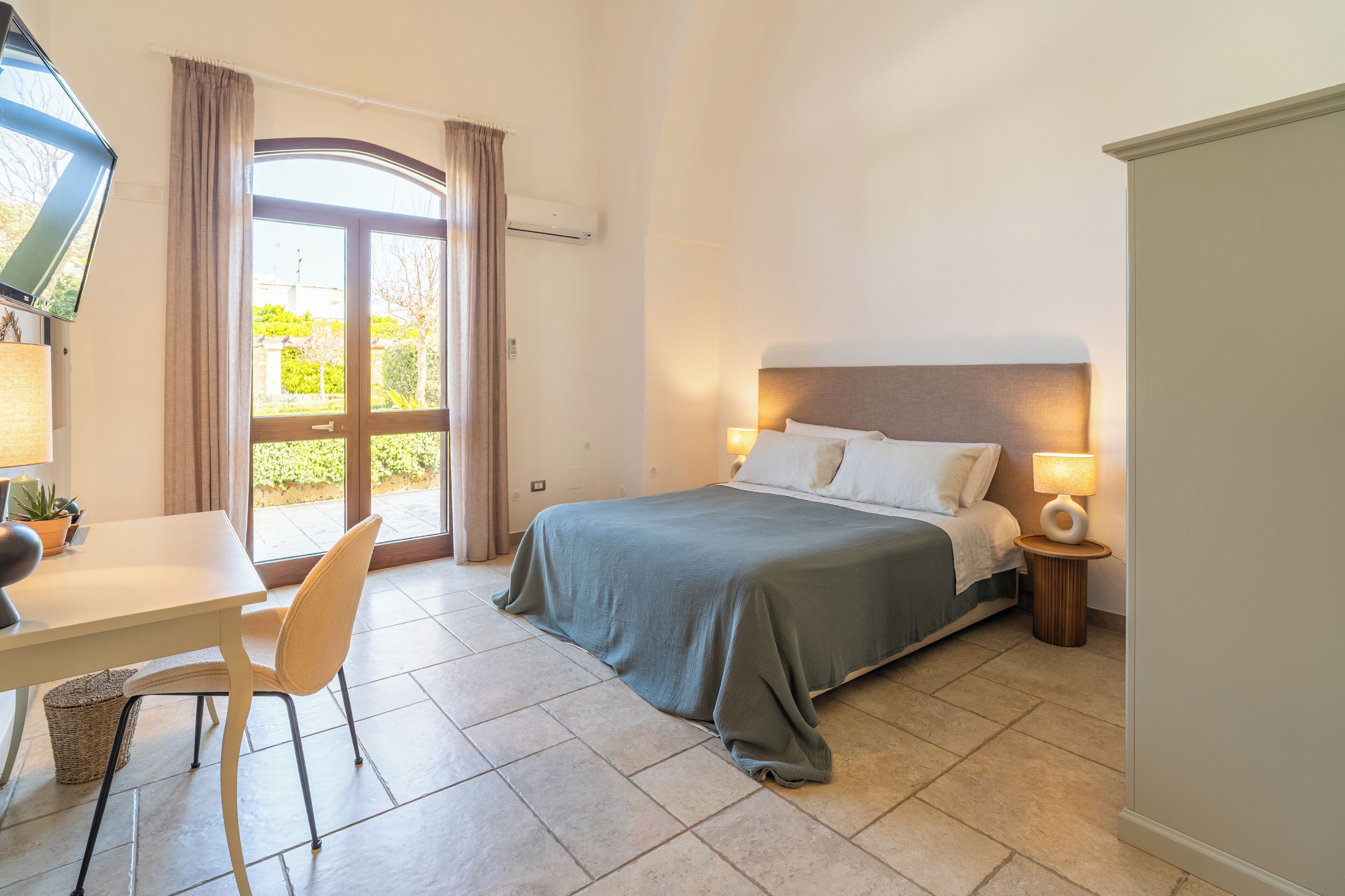 Habitación de hotel con cama, escritorio, silla, televisor y ventana con cortinas.