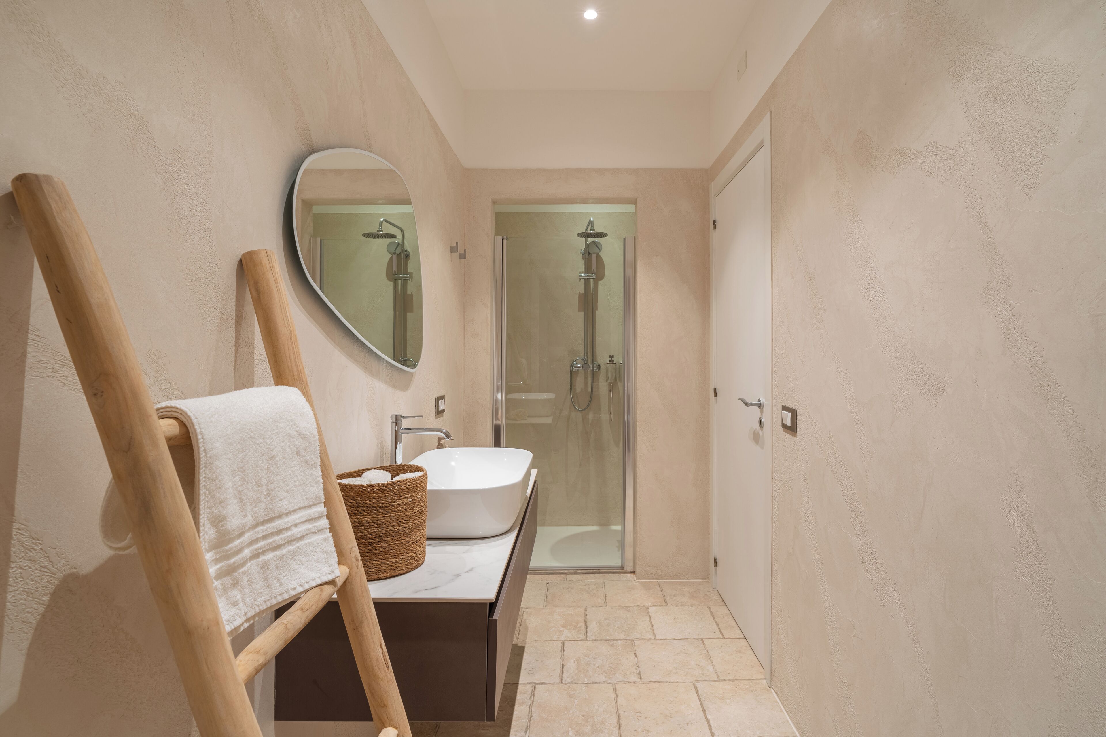 A bathroom with a wooden chair, a round mirror, a white sink, and a glass-enclosed shower.