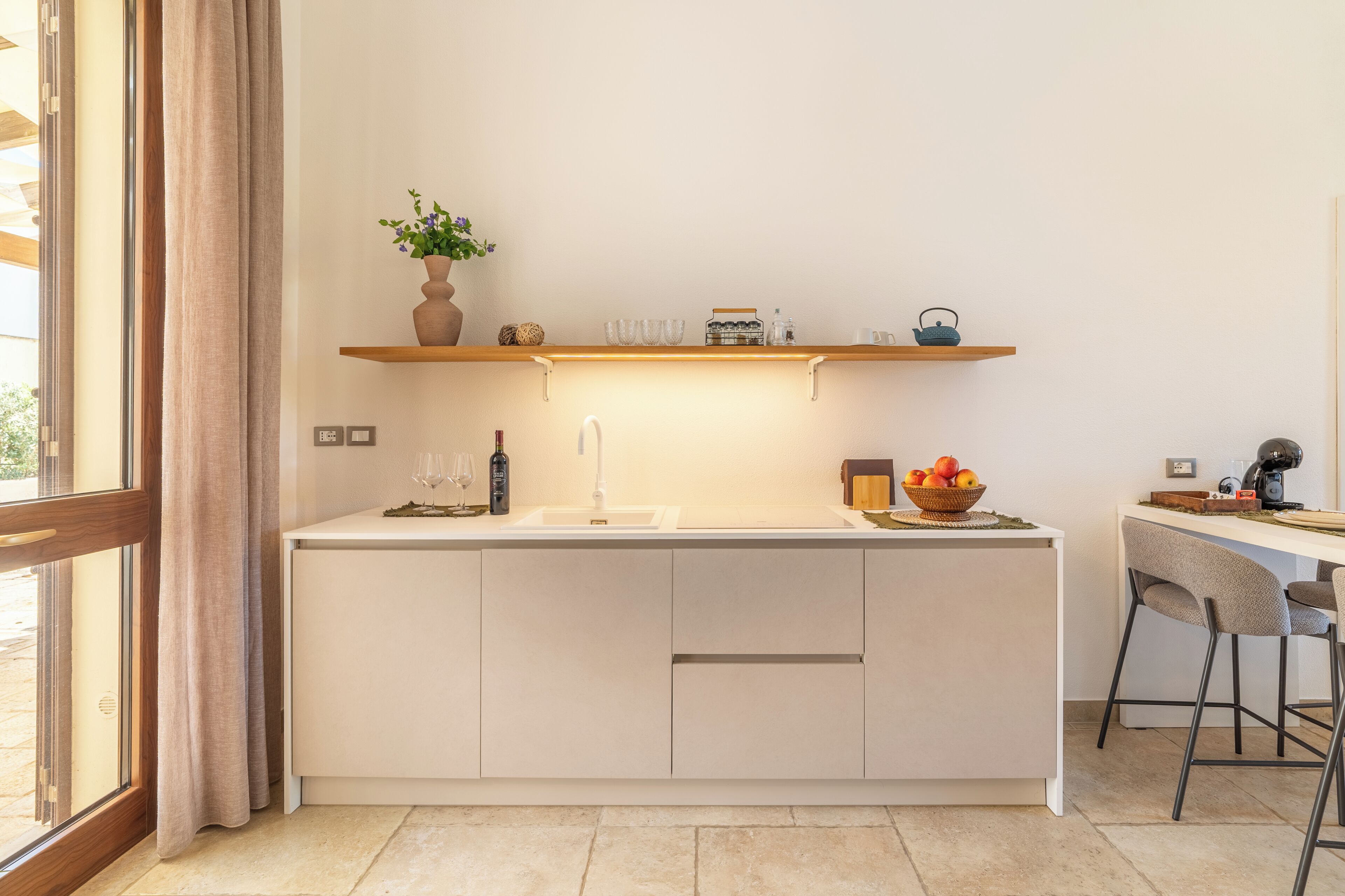 A modern kitchen with a white countertop, a sink, a shelf with decorative items, and a window with curtains.