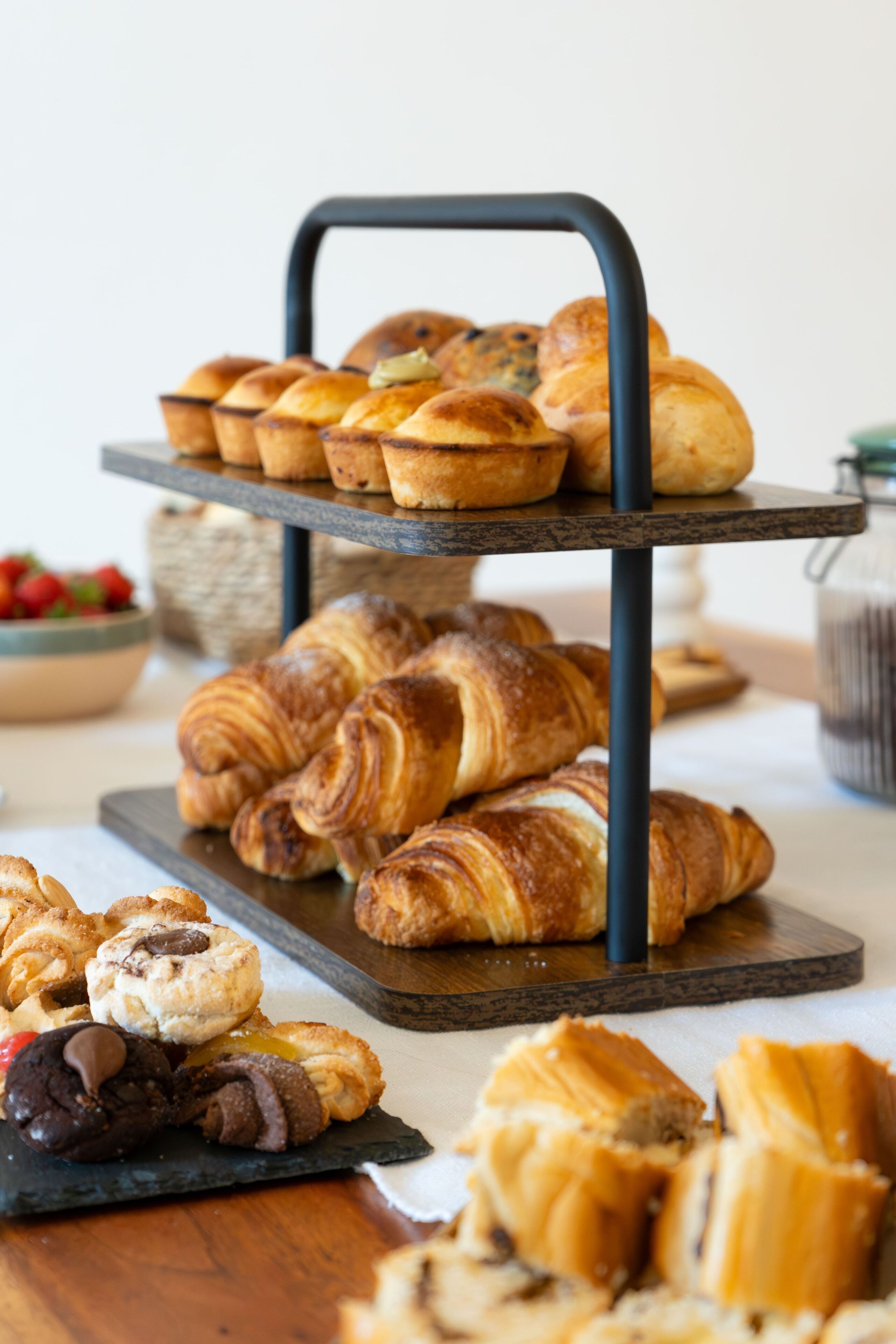 A tiered serving tray with assorted pastries and a bowl of fruit in the background.