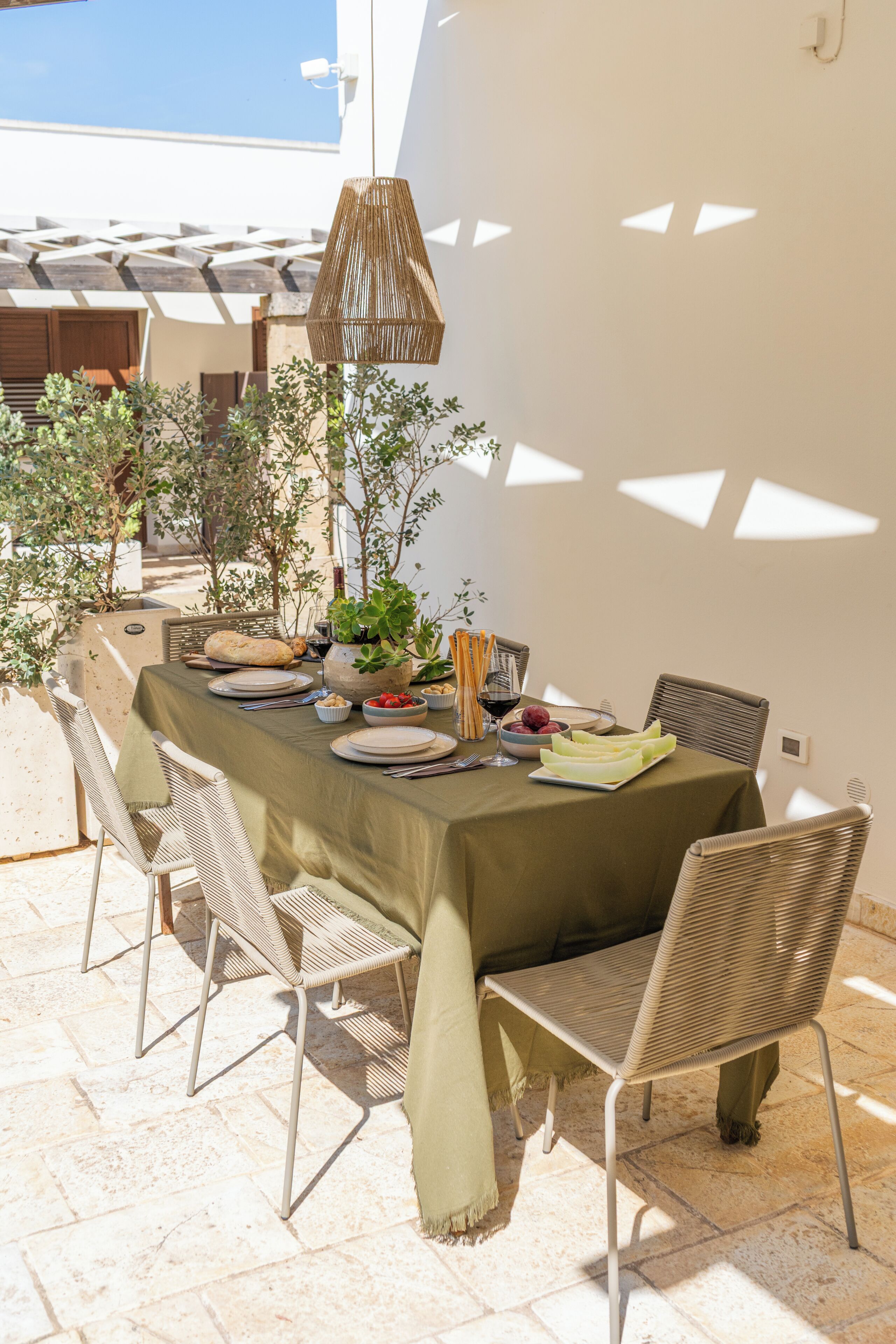 An outdoor dining area with a table set for four, surrounded by wicker chairs, with a pergola overhead and potted plants.