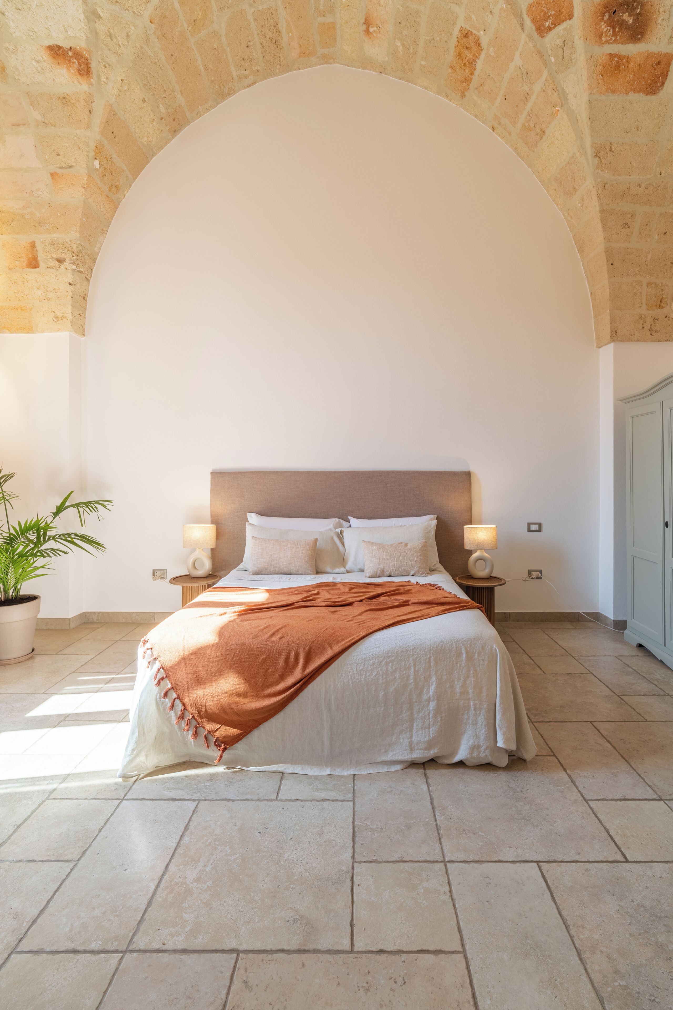Un dormitorio con una cama grande, dos lámparas de noche, una planta en maceta y un arco de pared de piedra.