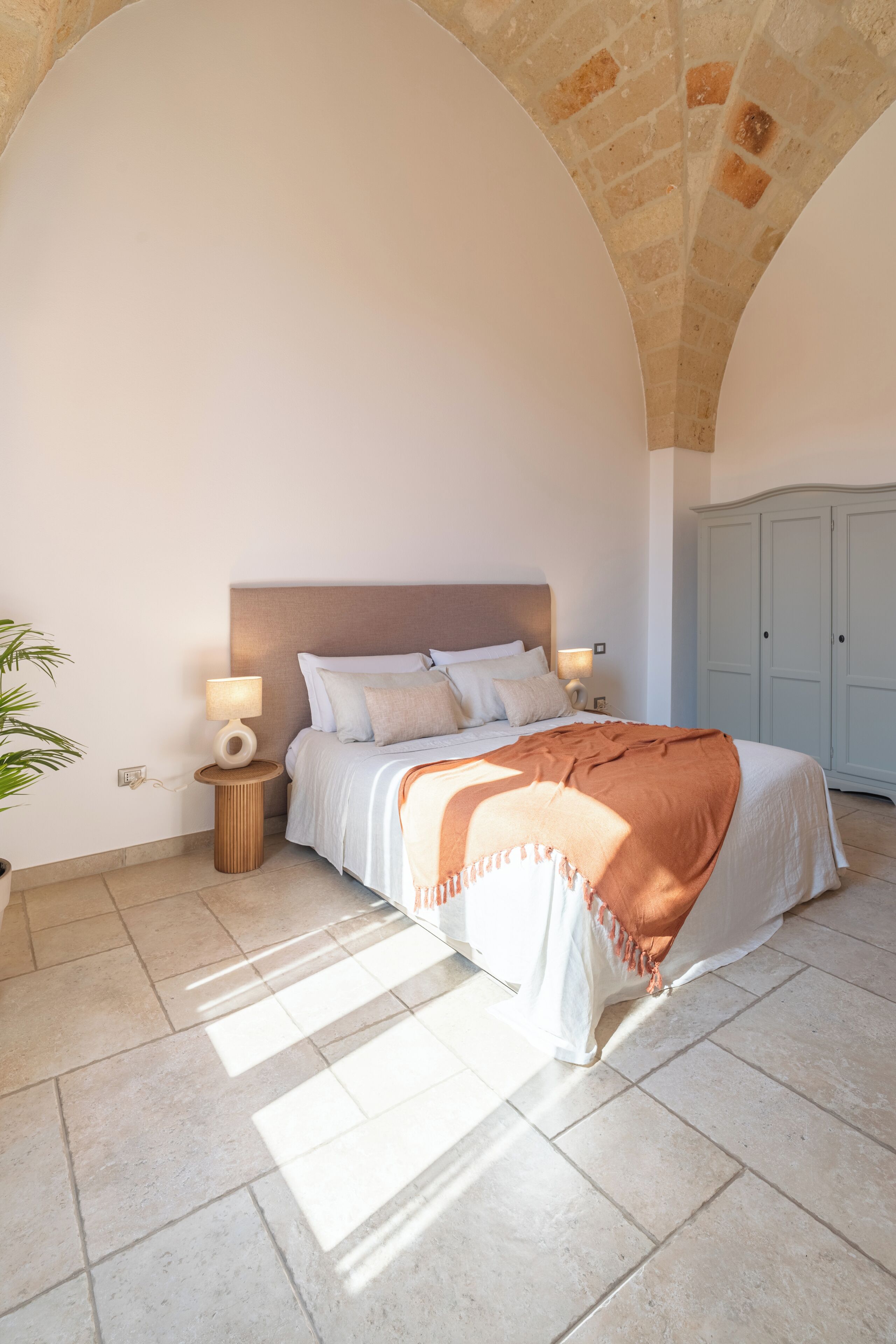 A bedroom with a bed, bedside lamps, a nightstand, a wardrobe, and a potted plant.