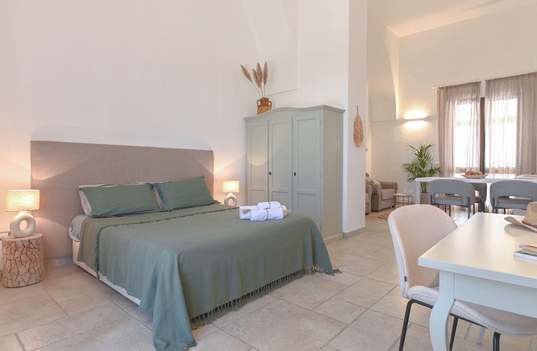 A bedroom with a bed, bedside lamps, a wardrobe, and a dining area with chairs and a table.