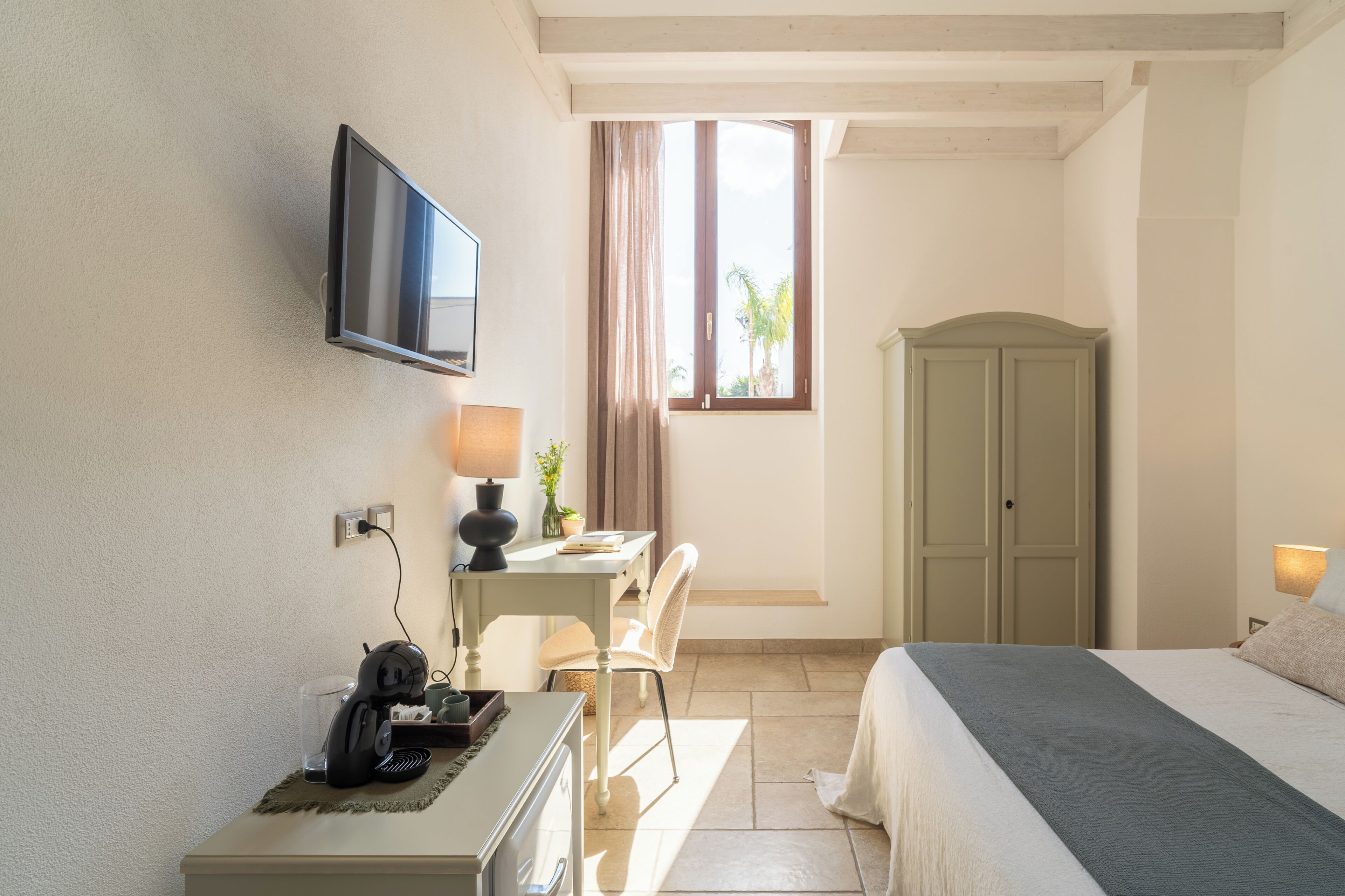 Une chambre d’hôtel comprenant un lit, un bureau, une chaise, une télévision, une armoire et une fenêtre avec des rideaux.
