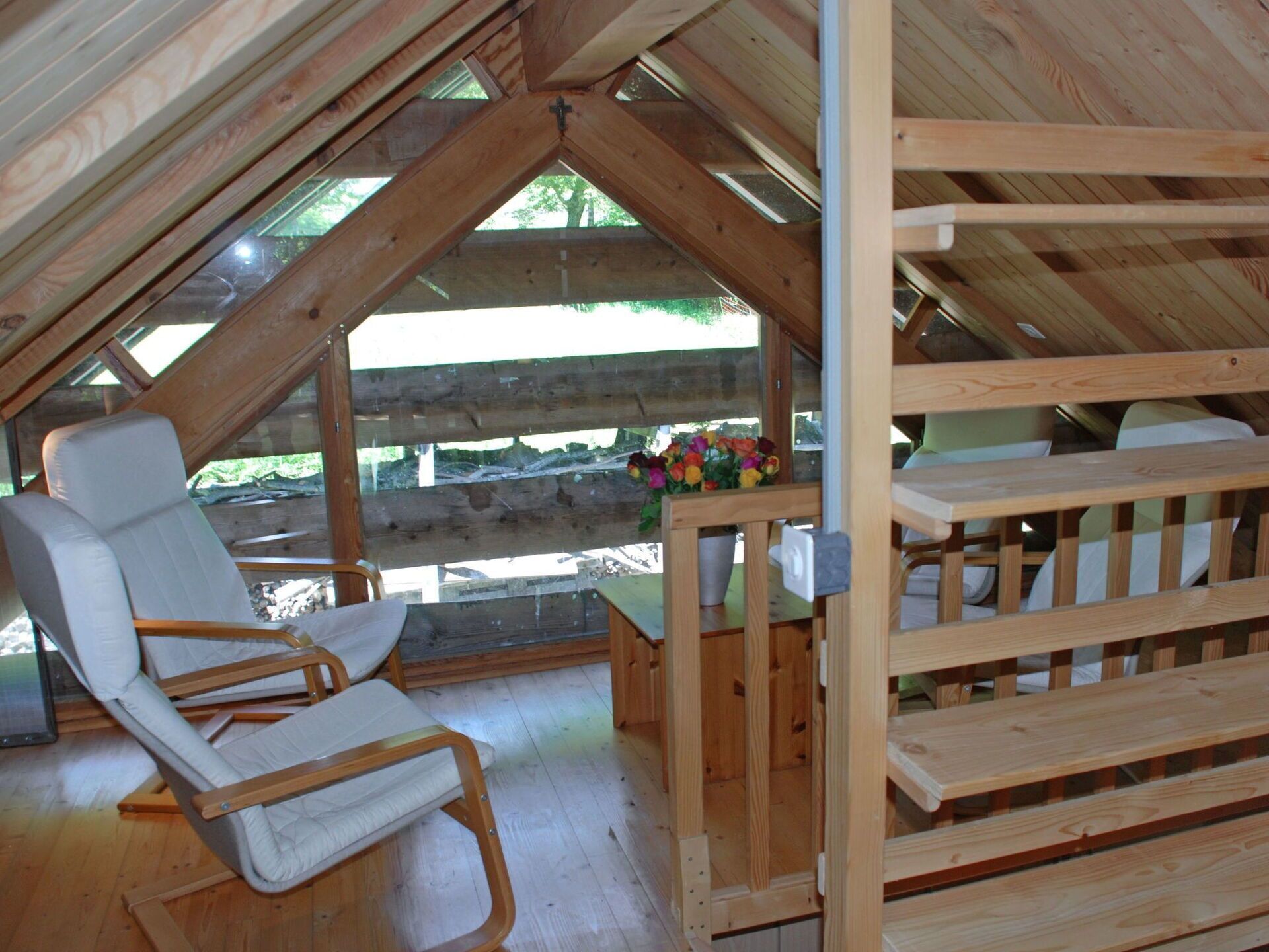 A wooden loft bedroom with a sloped ceiling, a wooden bench, a cushioned chair, and a small table with a flower vase.