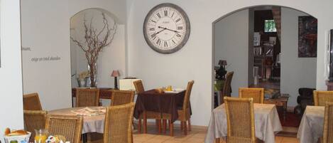 A dining area with wicker chairs, tables, a large wall clock, and a bookshelf in the background.
