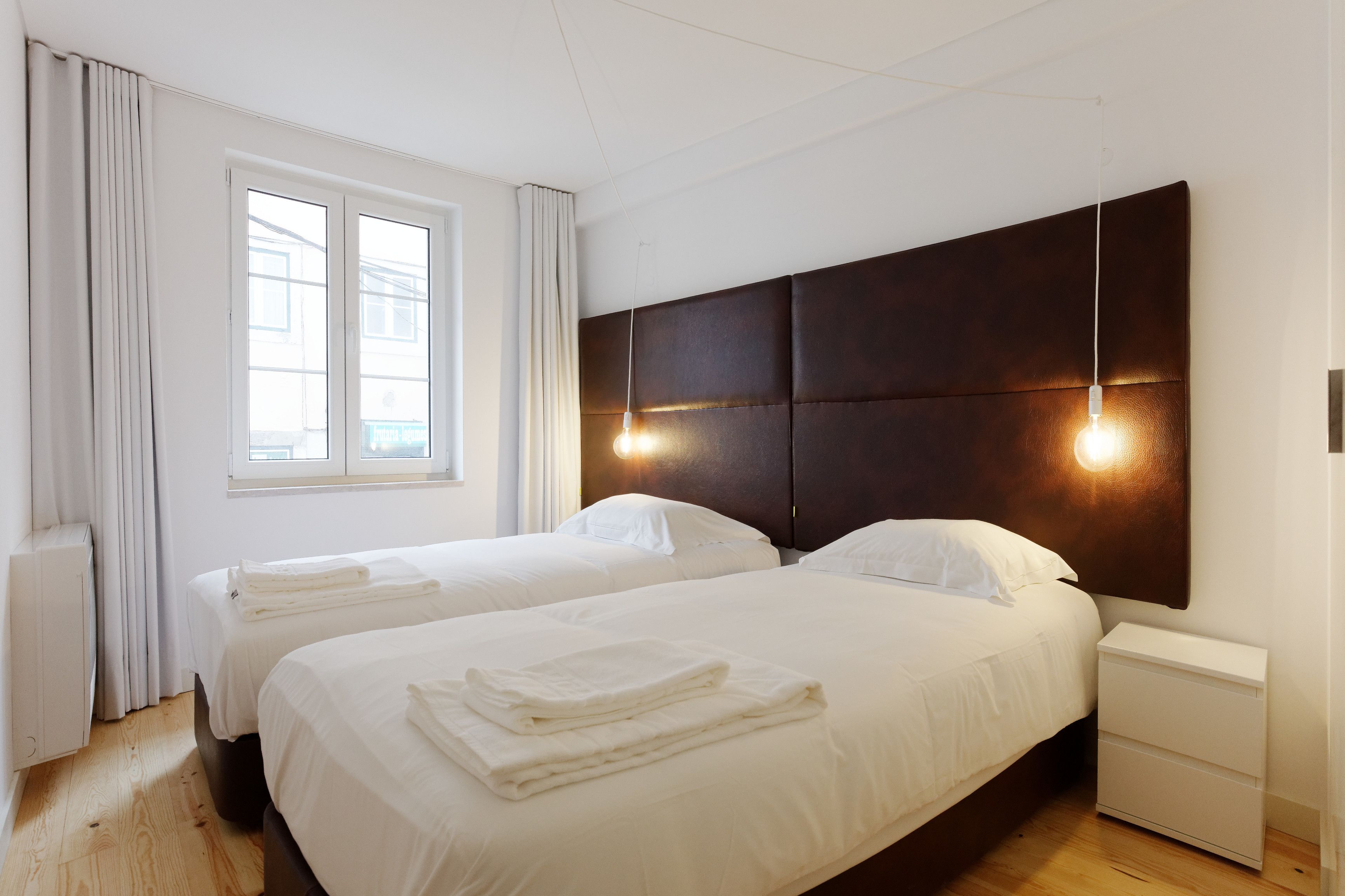 Ein Hotelzimmer mit zwei Betten, einem großen Fenster, einem dunklen Kopfteil und einem Nachttisch mit Lampe.
