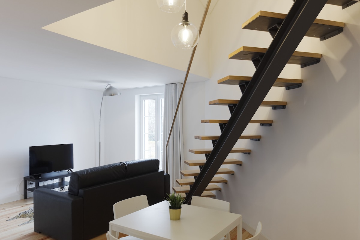 A modern living room with a black sofa, a white dining table, and a staircase with wooden steps.