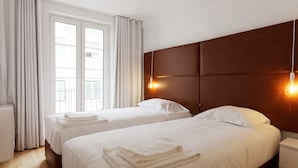Quarto de hotel com duas camas, chão de madeira, janela grande com cortinas e cabeceira marrom com luzes fixadas na parede.