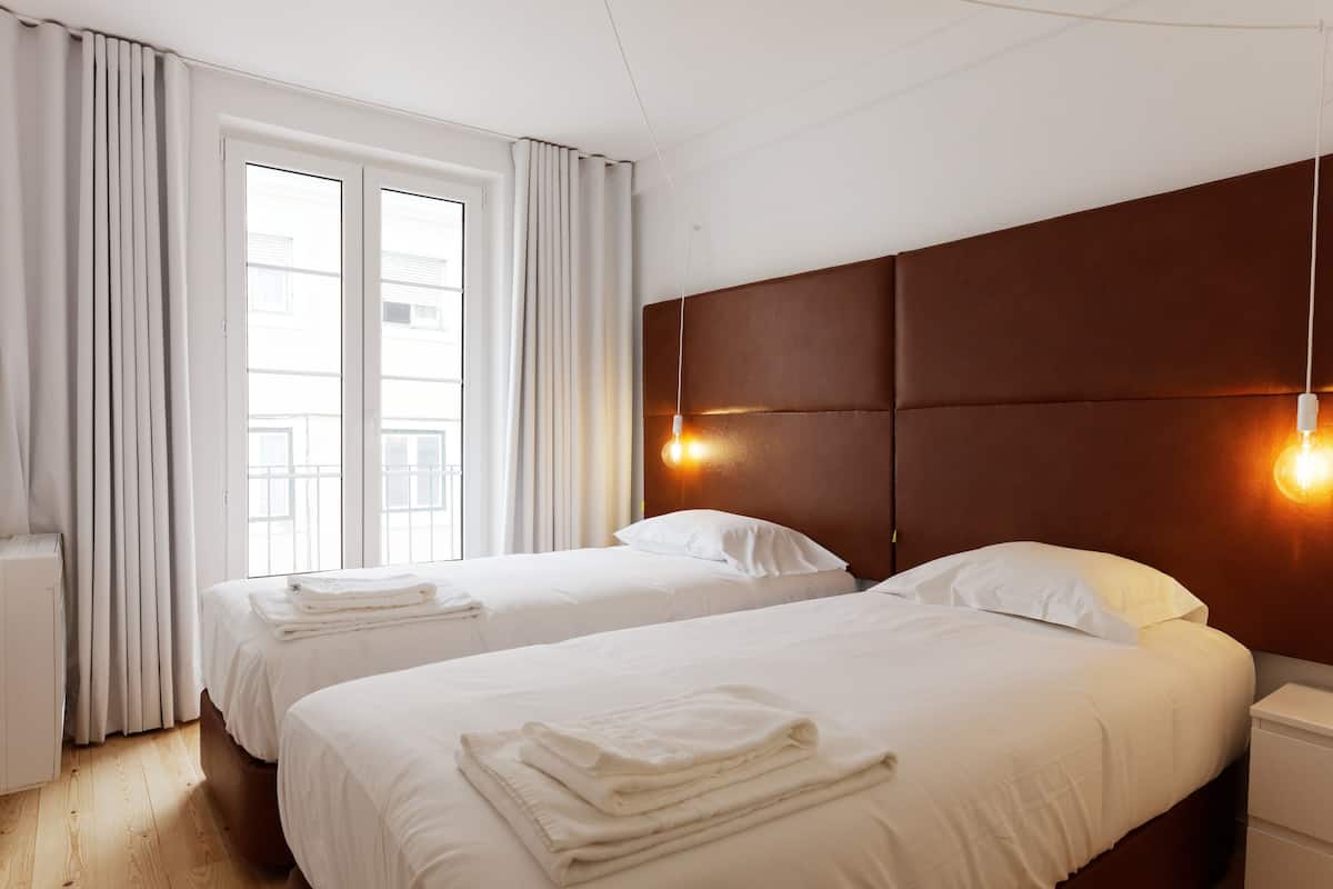 A hotel room with two beds, a wooden floor, a large window with curtains, and a brown headboard with wall-mounted lights.