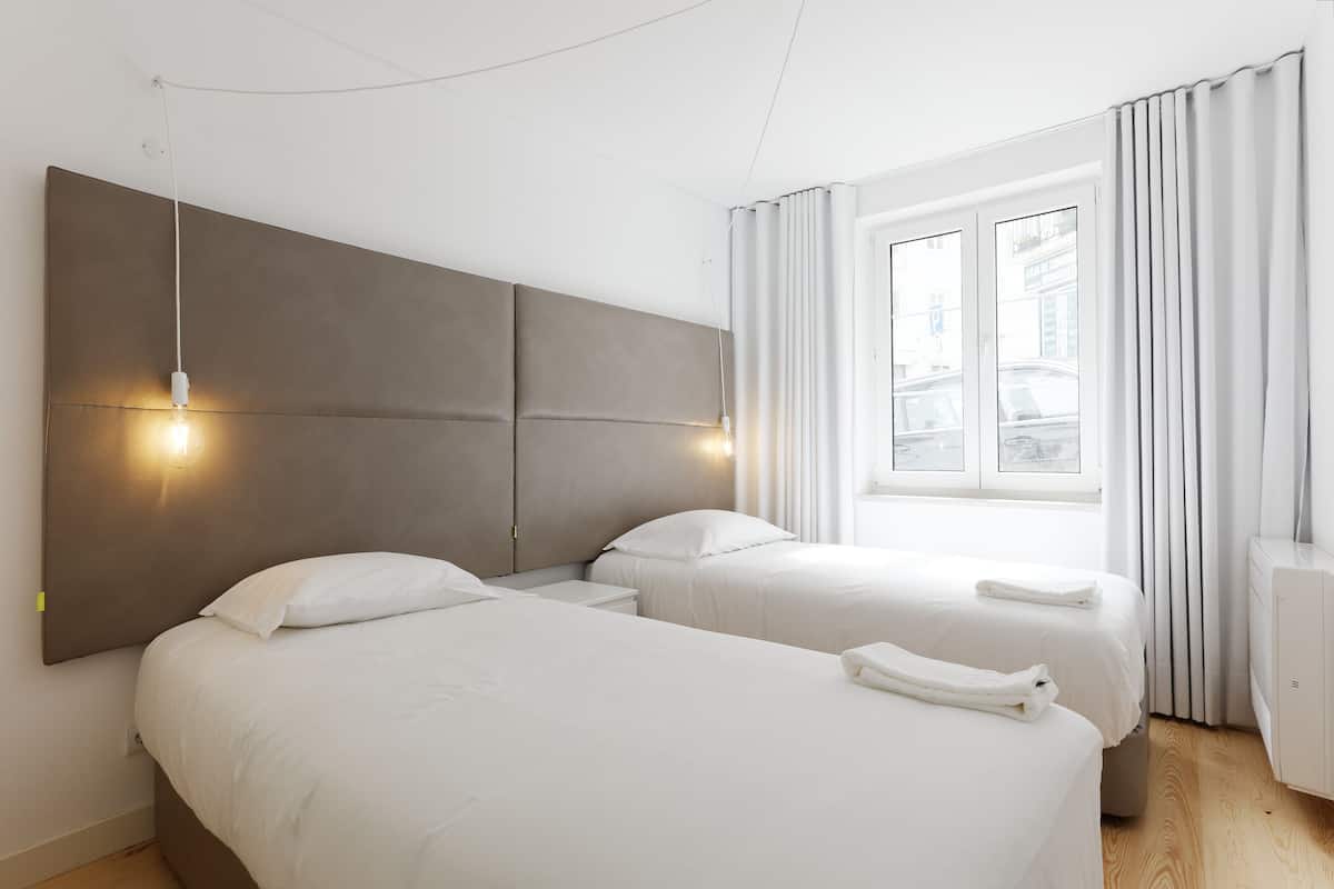 A hotel room with two beds, a large headboard, a window with curtains, and a bedside table.