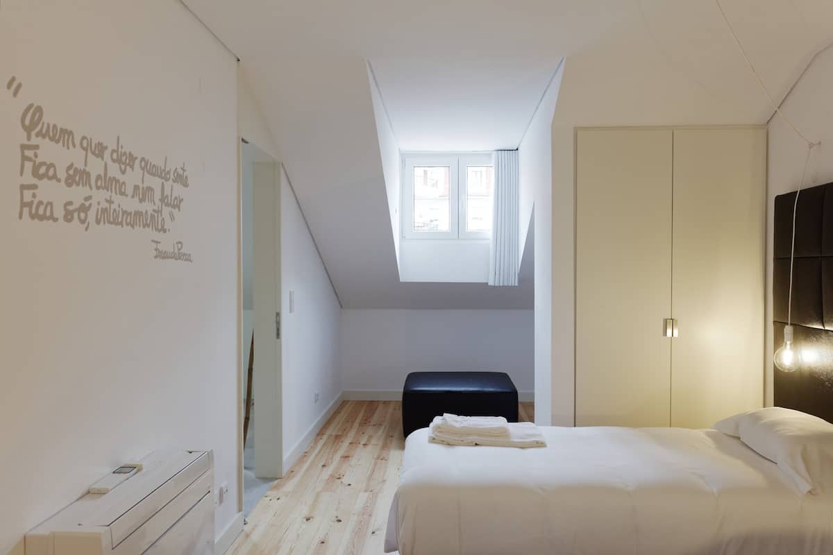 A bedroom with a loft-style ceiling, a bed with white bedding, a small bench, and a window with curtains.