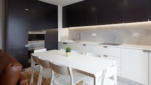 Uma cozinha moderna com armários escuros, bancada branca e área de refeições com mesa e cadeiras brancas.