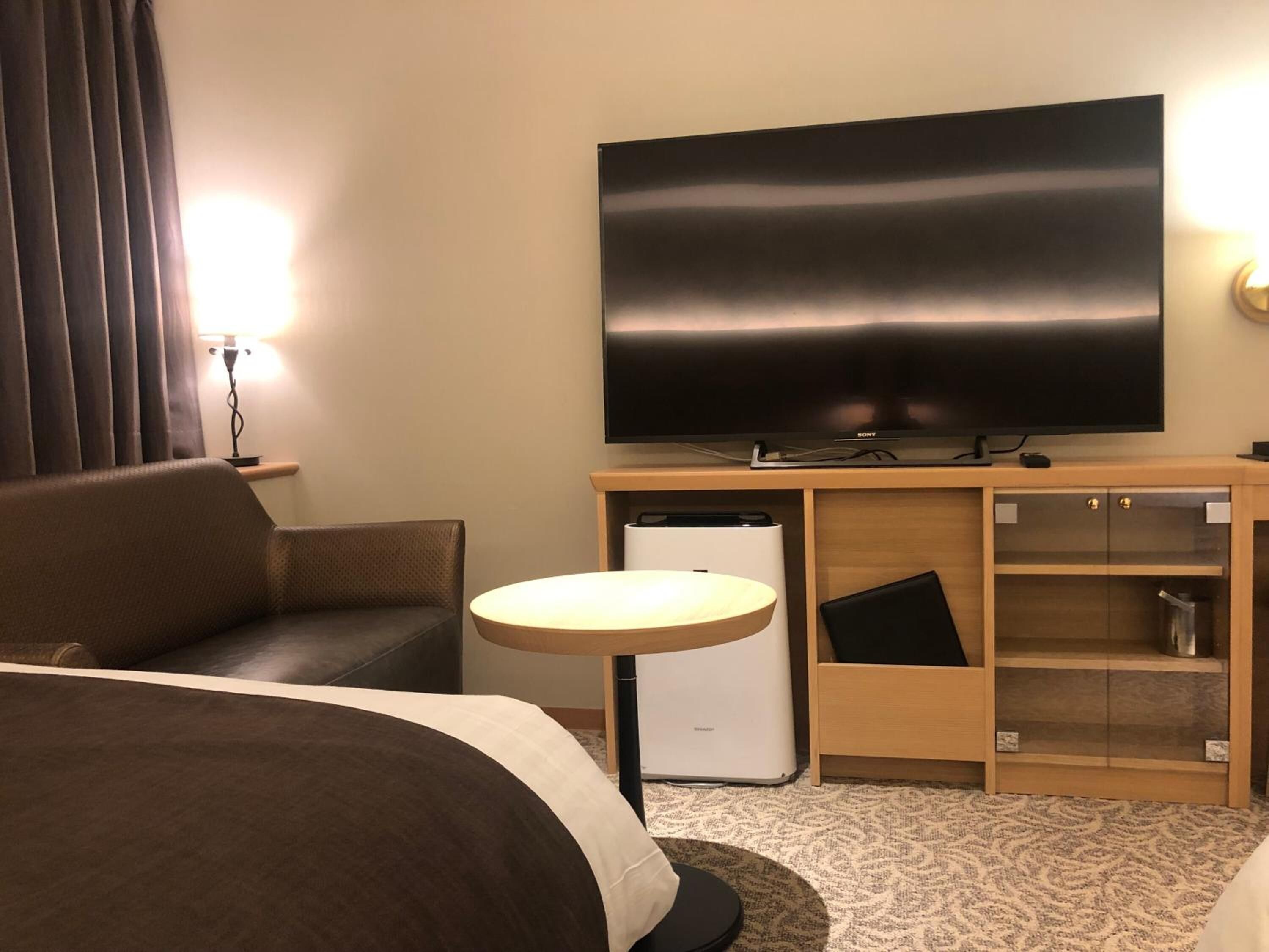 Quarto de hotel com uma televisão de tela plana grande, um sofá, uma mesa redonda pequena, um mini-refrigerador e um armário de madeira com portas de vidro.