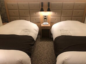 Quarto de hotel com duas camas, uma mesinha de cabeceira com telefone e cabeceira com luz.