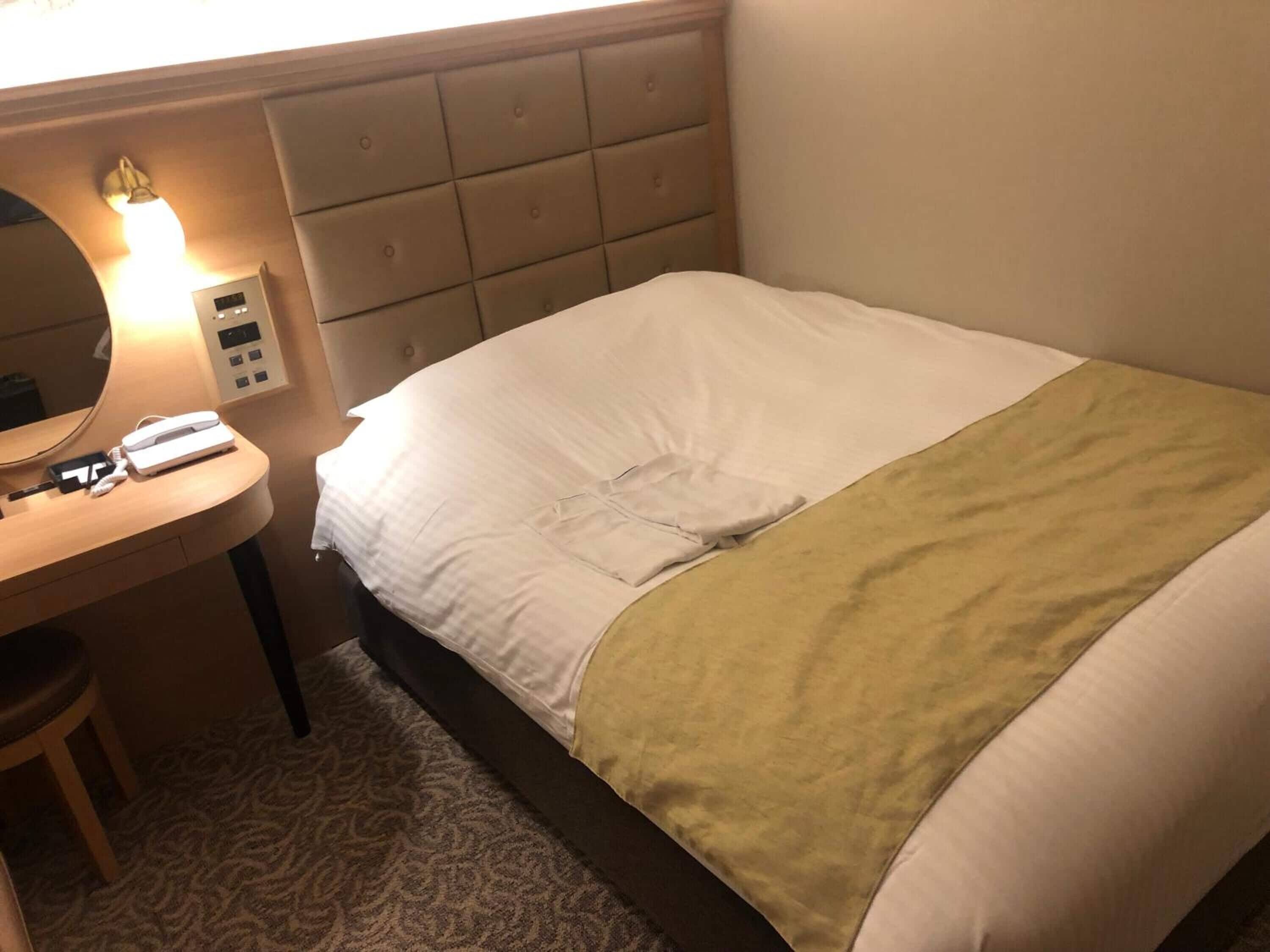 Habitación de hotel con una cama, una mesita de noche con lámpara, un teléfono y un panel de control.