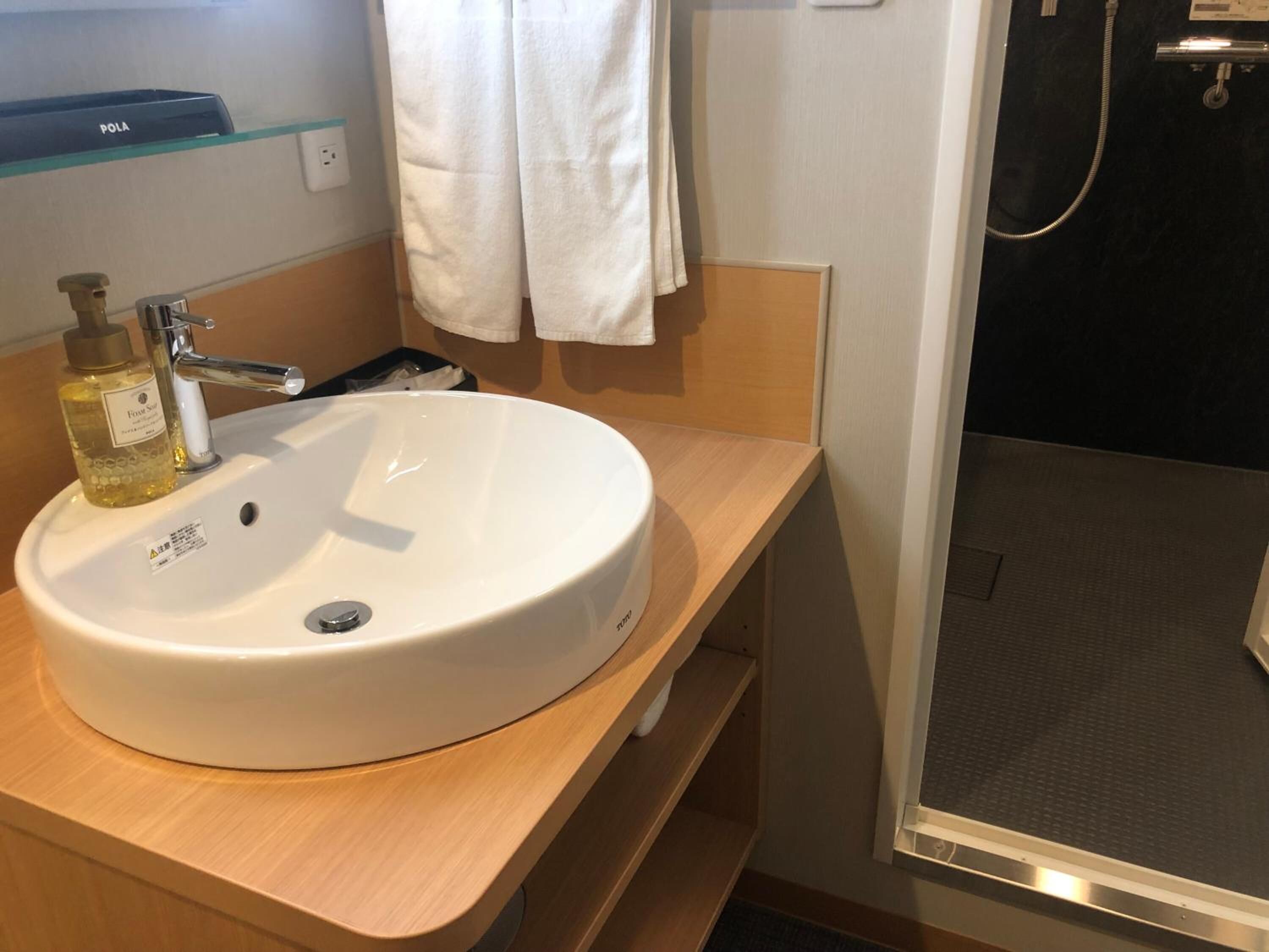 Un baño con lavamanos blanco, una botella de jabón, un espejo y zona de ducha.