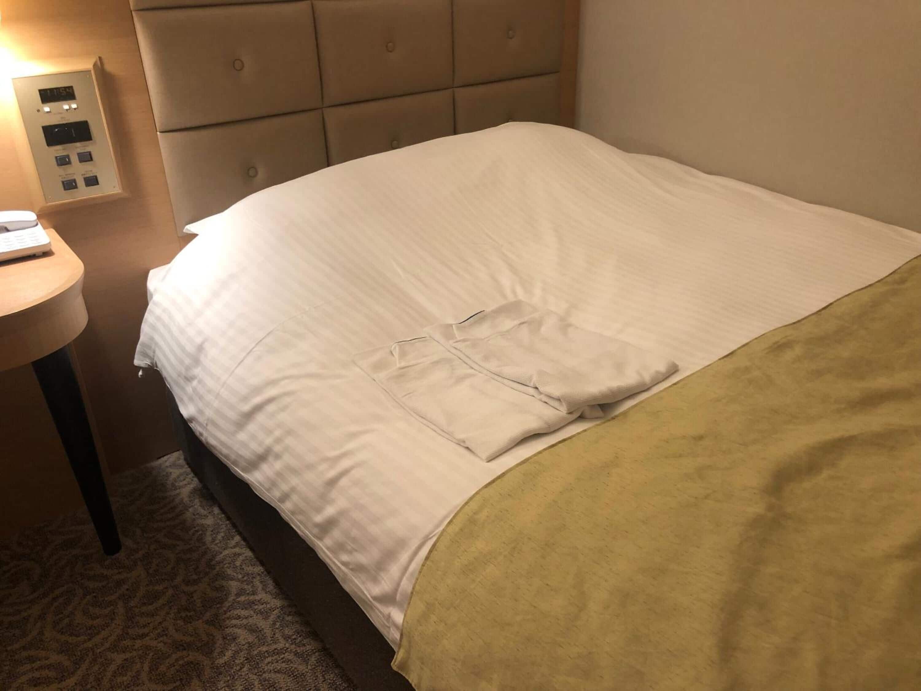 A neatly made bed with a white comforter and a yellow blanket, a bedside table with a lamp, and a control panel on the wall.