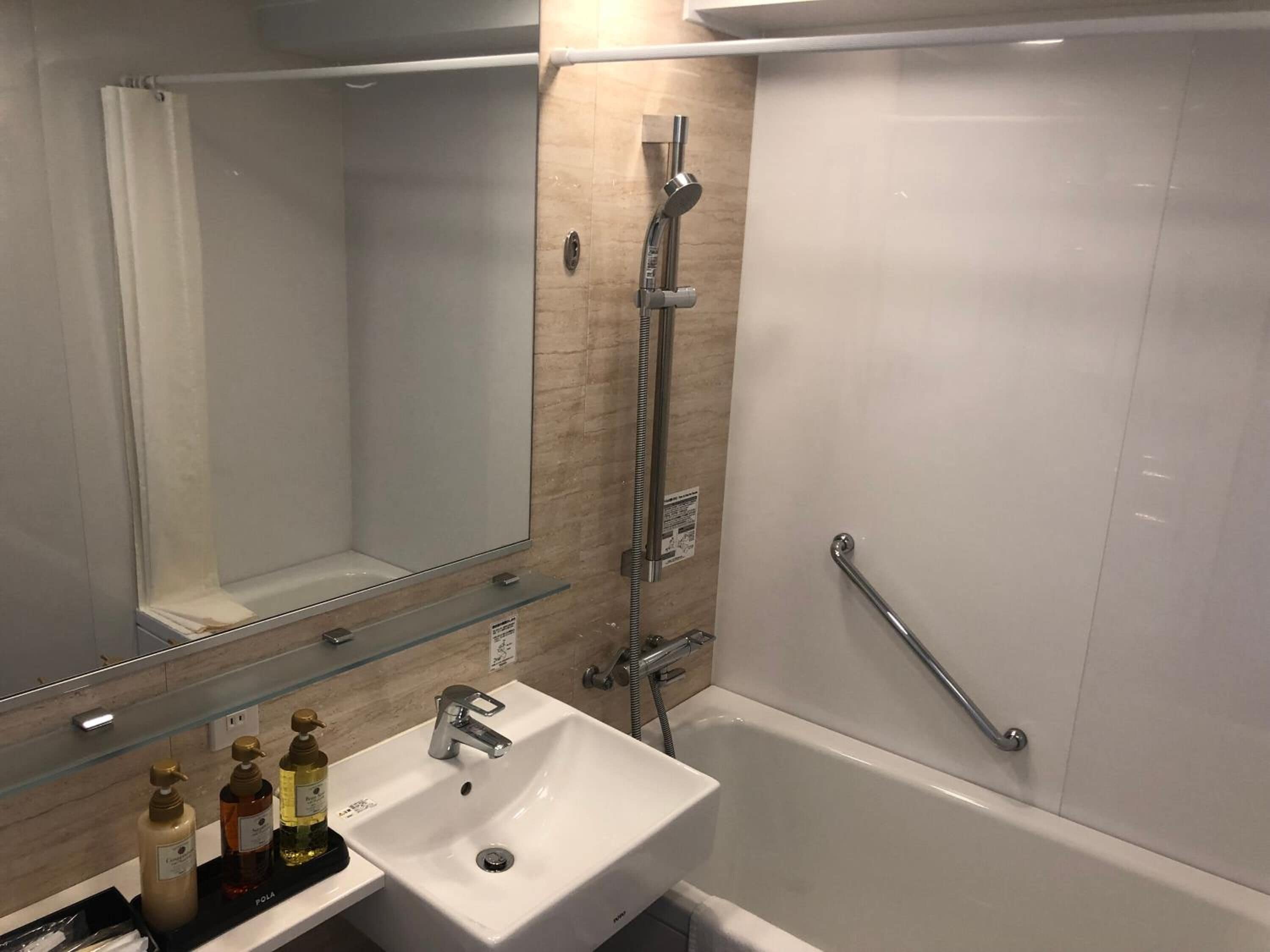 A modern bathroom with a white sink, a glass shelf with toiletries, a shower with a handheld showerhead, and a grab bar.