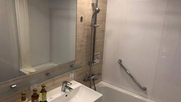 A modern bathroom with a white sink, a glass shelf with toiletries, a shower with a handheld showerhead, and a grab bar.