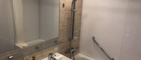 A modern bathroom with a white sink, a glass shelf with toiletries, a shower with a handheld showerhead, and a grab bar.